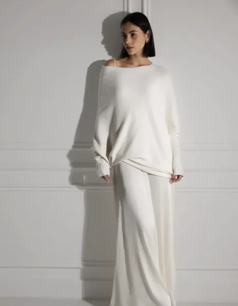 Woman in white off-shoulder sweater and wide-leg pants posing against a white wall