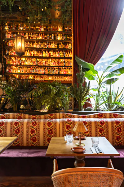 Cozy restaurant booth with patterned upholstery, wooden table set with glasses and lamp, glowing bar shelves with bottles, and large plants by a window with red curtains