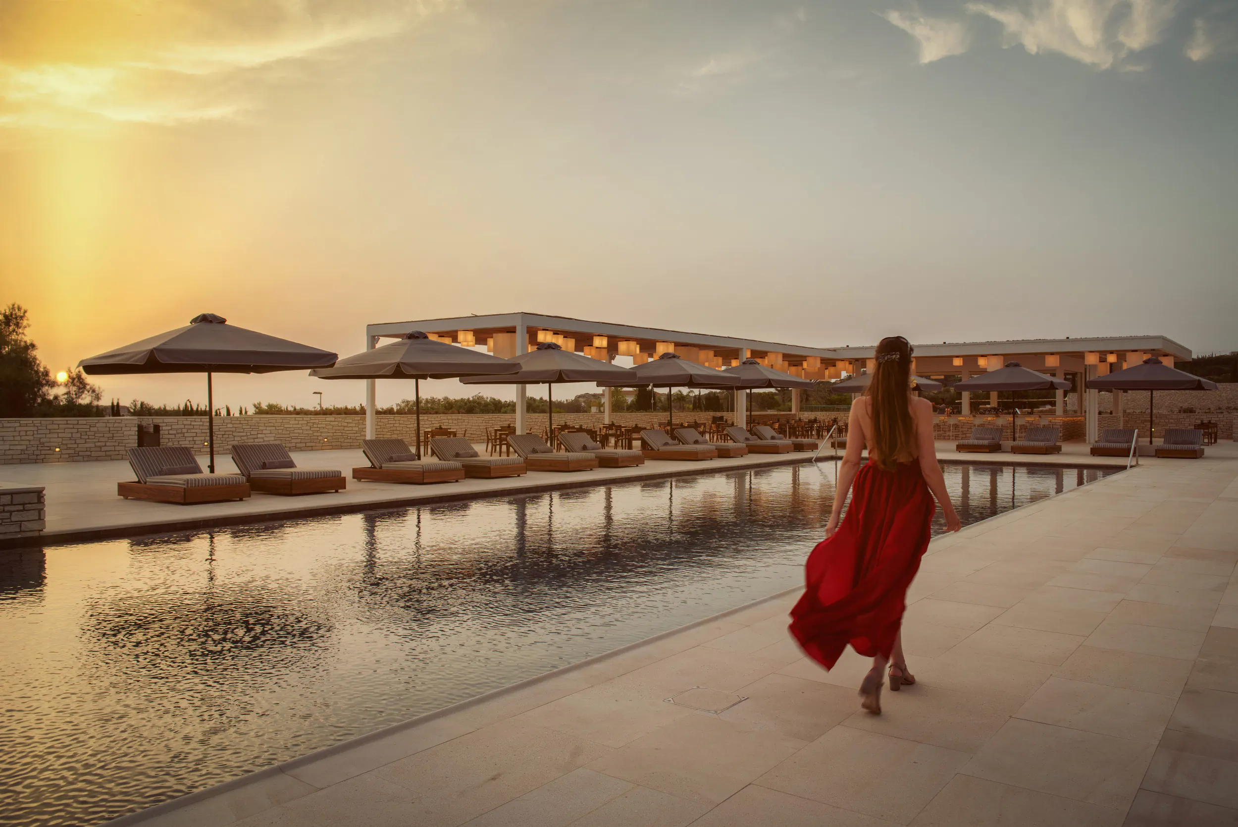 Woman in flowing red dress walking by infinity pool at sunset with resort lounge chairs and umbrellas