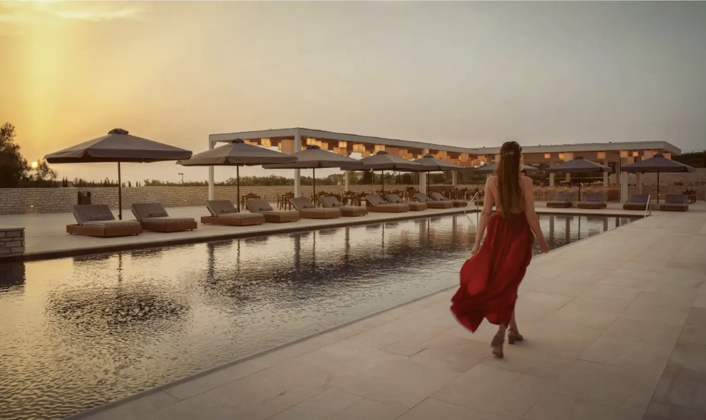 Woman in flowing red dress walking by infinity pool at sunset with lounge chairs and umbrellas