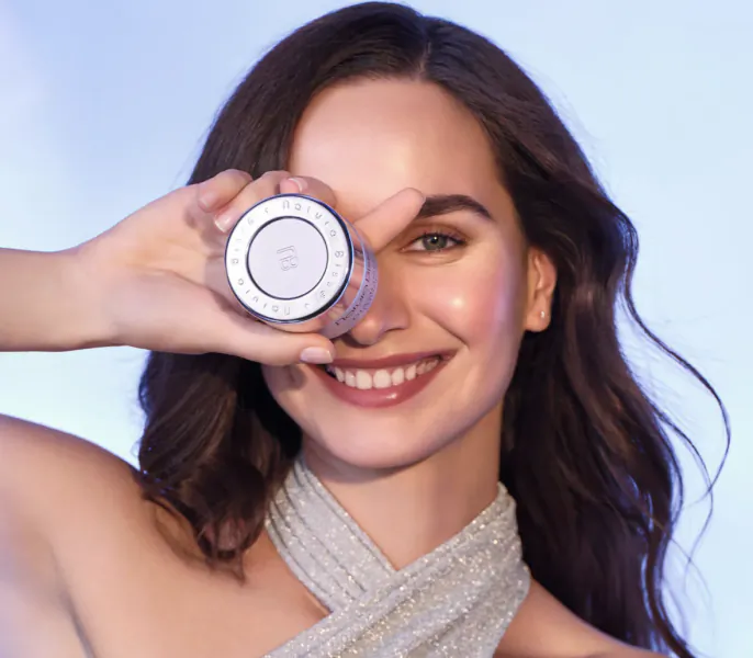 Smiling woman with brown hair and silver dress winking through round skincare jar labeled 'Smart energy for skin? Always'