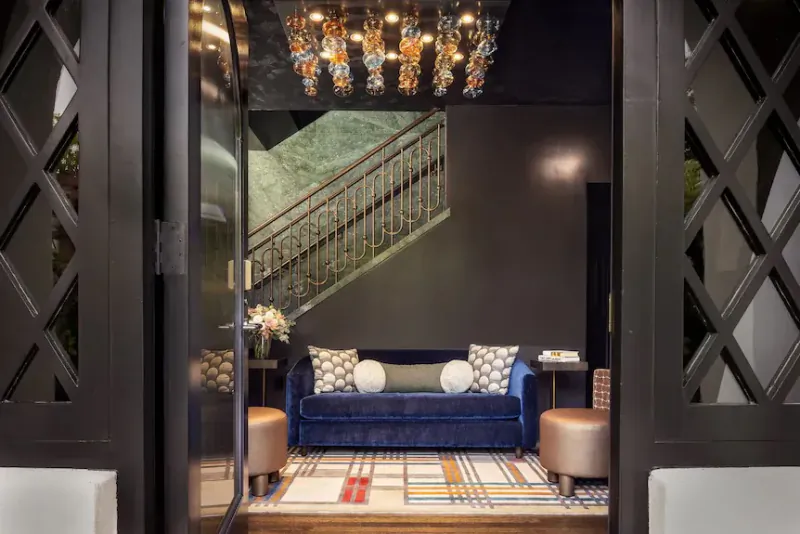 Elegant hotel lobby with blue velvet sofa, patterned rug, and ornate chandelier viewed through open dark doors