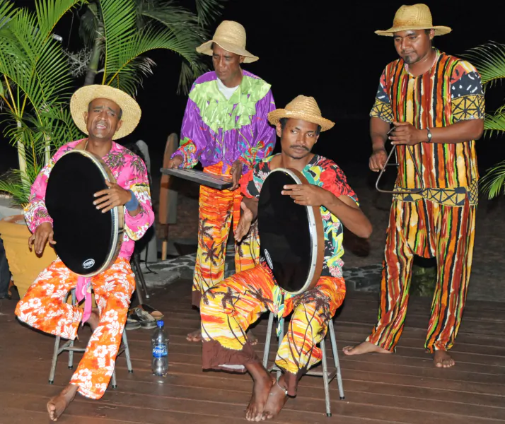 Four men in colorful patterned outfits and straw hats play drums on a wooden deck at night
