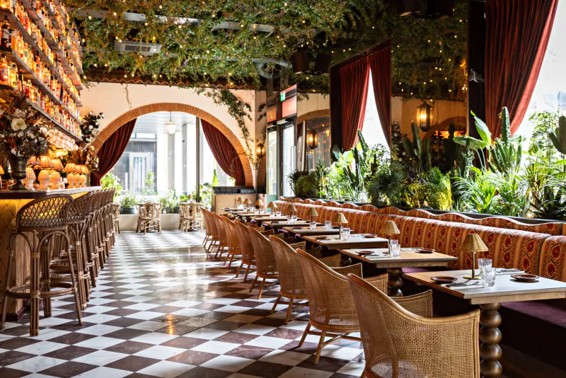 Elegant restaurant interior with rows of tables, woven chairs, lush plants, and a long bar under arched ceilings