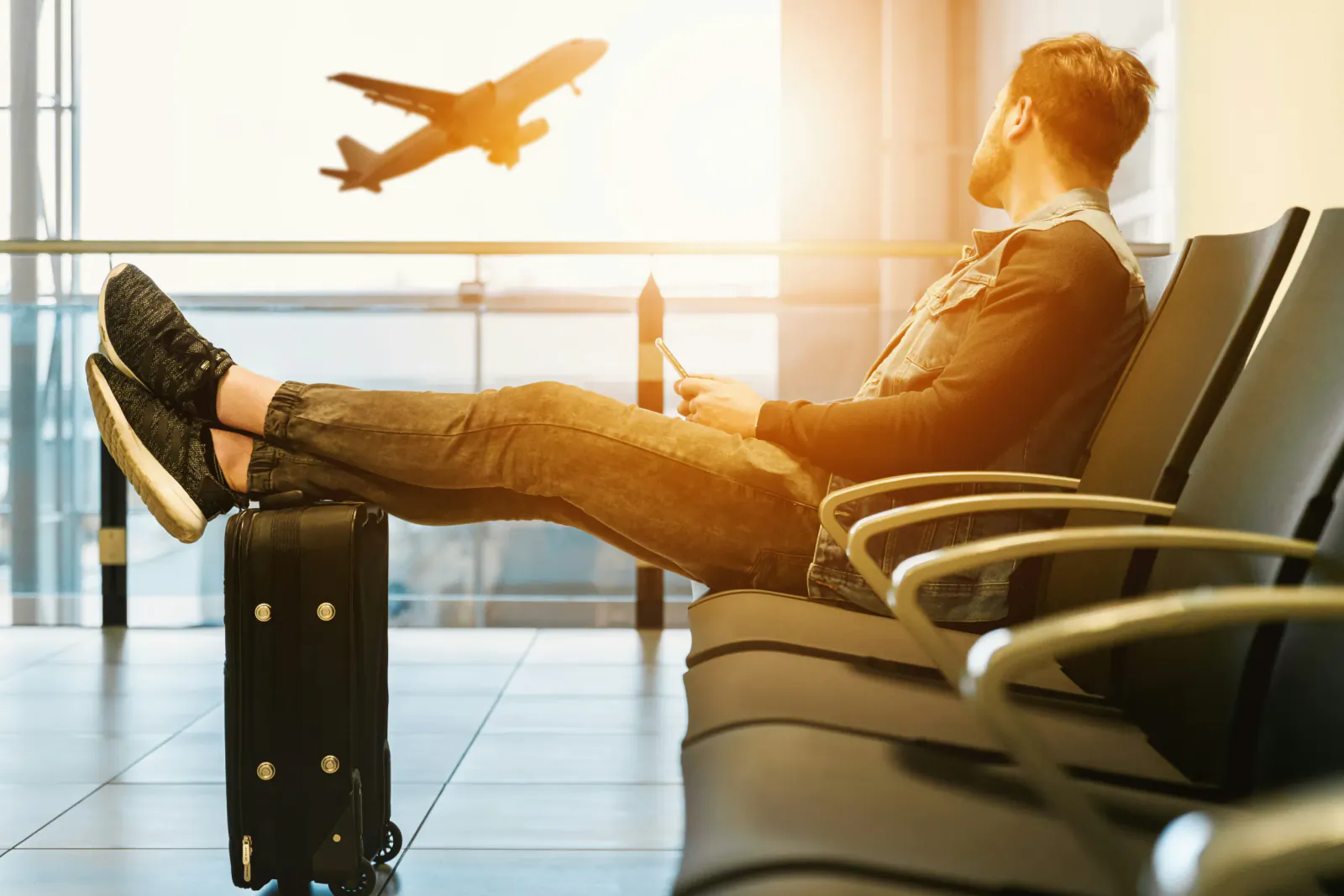 Man relaxing in airport lounge with feet on suitcase, checking phone as plane flies by window