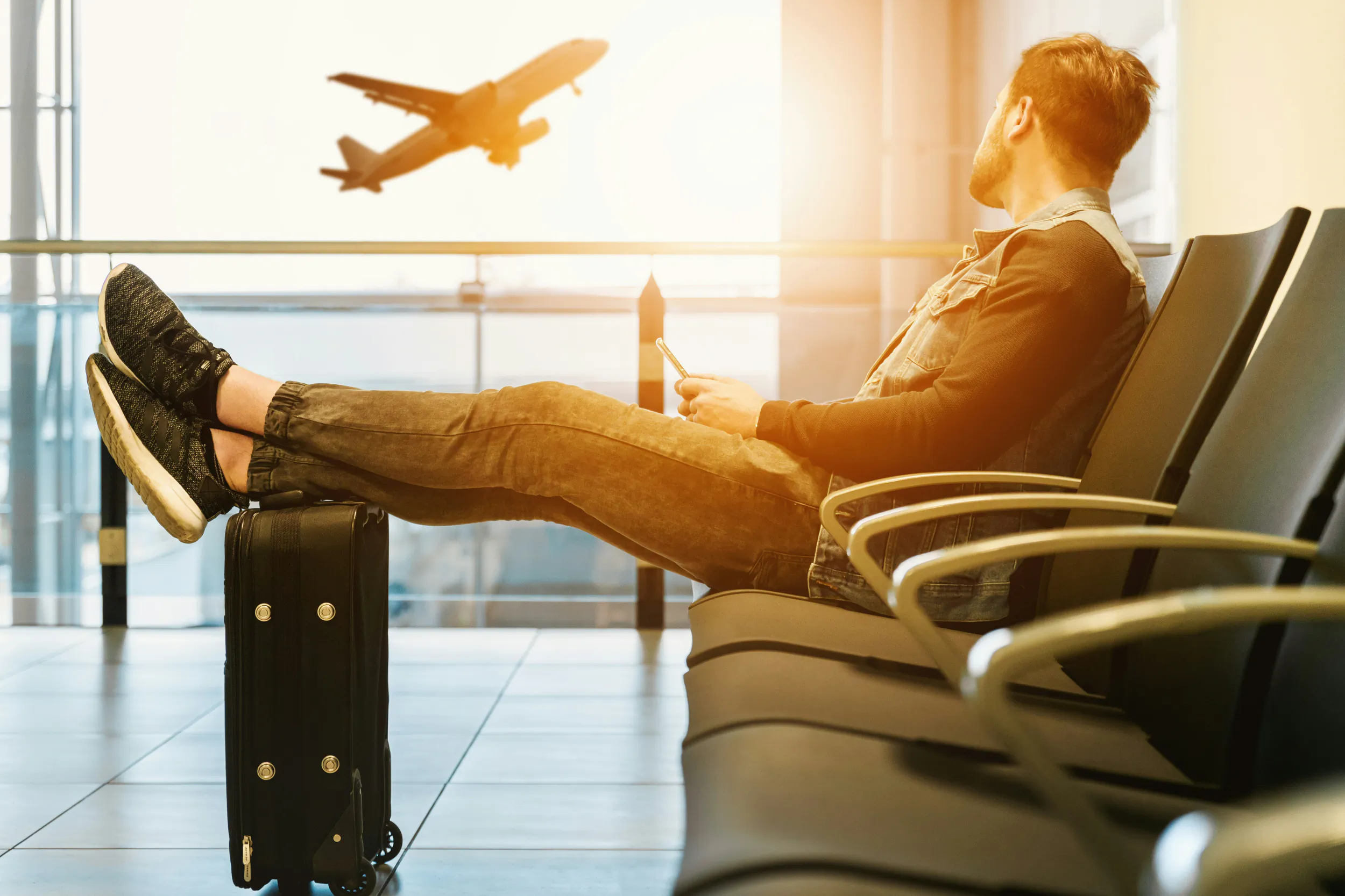 Man relaxing in airport lounge with feet on suitcase, checking phone as plane flies by window
