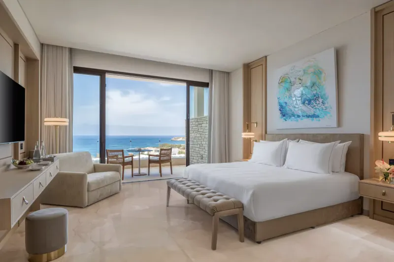 Luxurious hotel suite with king bed, seating area, and balcony overlooking Akamas sea and cliffs at Cap St Georges Resort.