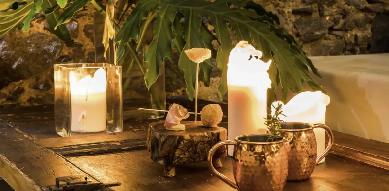 Lit candles, copper mugs, and natural decor on a wooden table with green leaves
