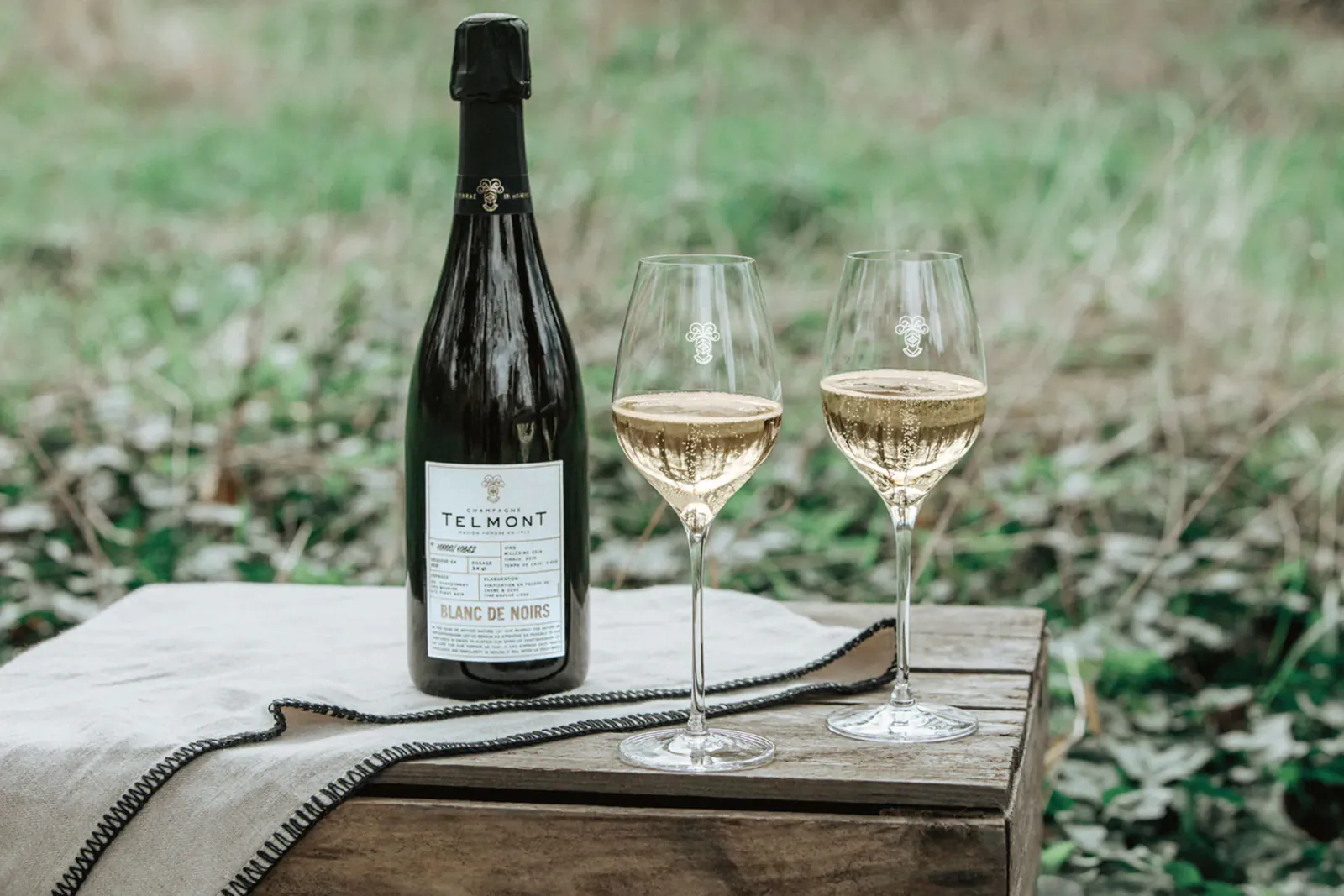 Bottle of Telmont Blanc de Noirs champagne with two glasses of sparkling wine on a wooden table outdoors