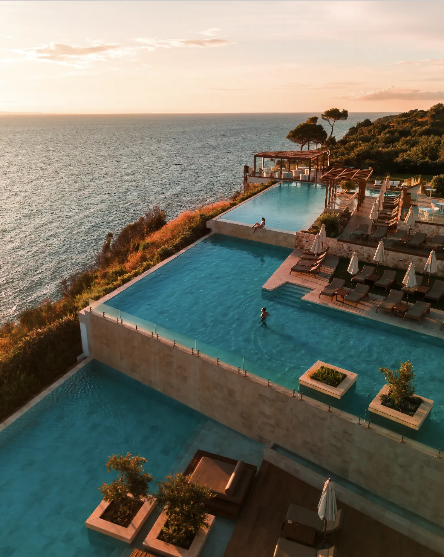 Aerial view of infinity pool overlooking ocean at sunset, with loungers, umbrellas, and swimmers.