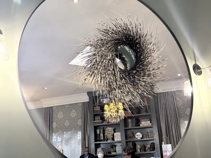 Round mirror reflecting a spiky silver wall sculpture, chandelier, and dark bookshelf in a hotel room