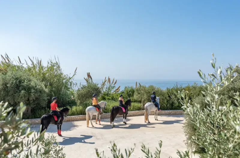 Four riders on horses in red, yellow, pink, and black at Cap St Georges Resort, overlooking sea with olive trees.