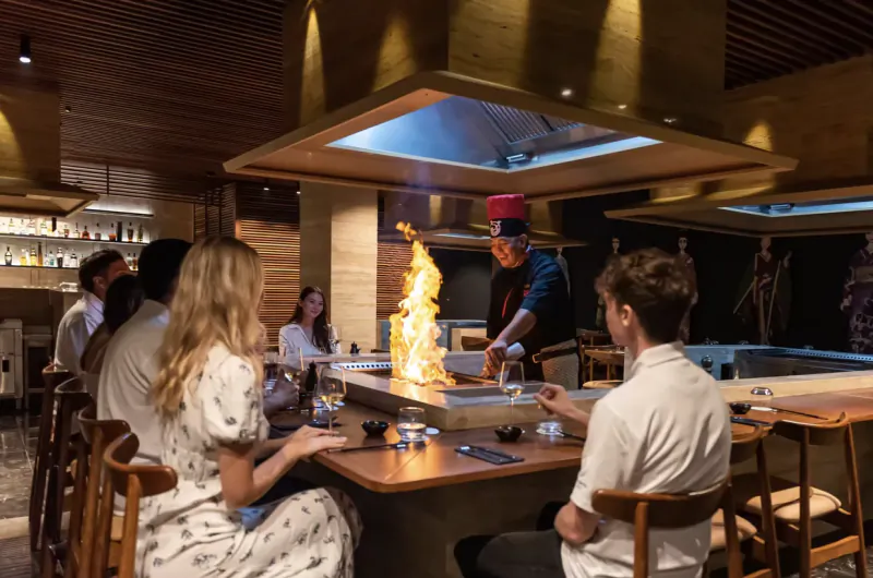 Chef in red headband performs teppanyaki with flames at hibachi grill, surrounded by diners at upscale restaurant