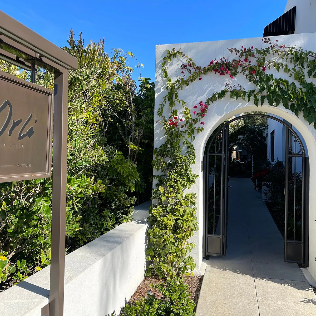 Hotel Orli sign beside a vine-covered white archway entrance with open black doors leading to a sunny pathway