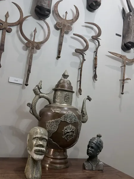 Ornate metal teapot with dragon spout and sculpted busts displayed below antique weapons on a white wall