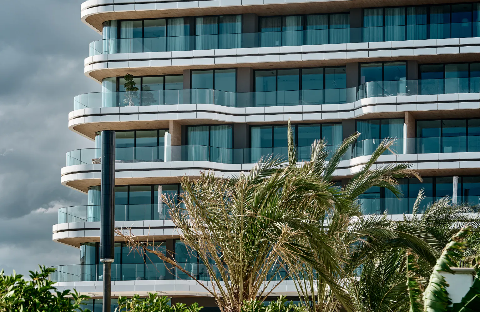 Modern curved high-rise apartment building with glass balconies and palm trees in foreground under cloudy sky