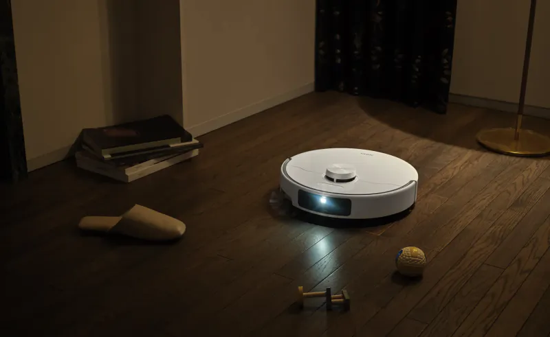 White robot vacuum cleaner with glowing light on dark wood floor, surrounded by books, a slipper, dumbbells, and a ball