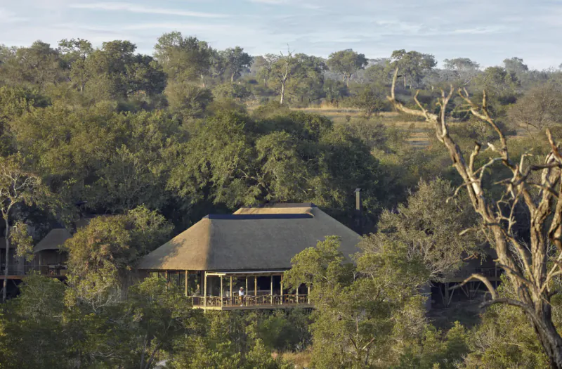 Thatched-roof lodge with deck nestled in lush African savanna bush, surrounded by trees under partly cloudy sky.