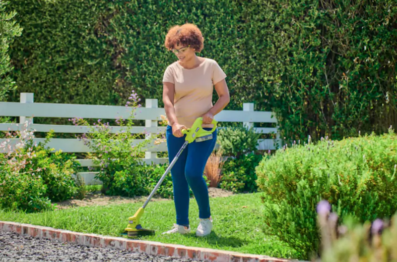 Woman using a green and yellow Ryobi string trimmer to edge the lawn in a sunny garden
