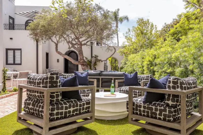 Modern outdoor patio with patterned armchairs, navy pillows, and a round white table on lush green lawn, surrounded by trees and white building