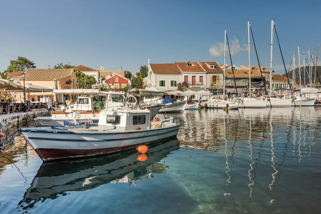 Serene harbor with white fishing boat, sailboats, and colorful Mediterranean village buildings under blue sky.