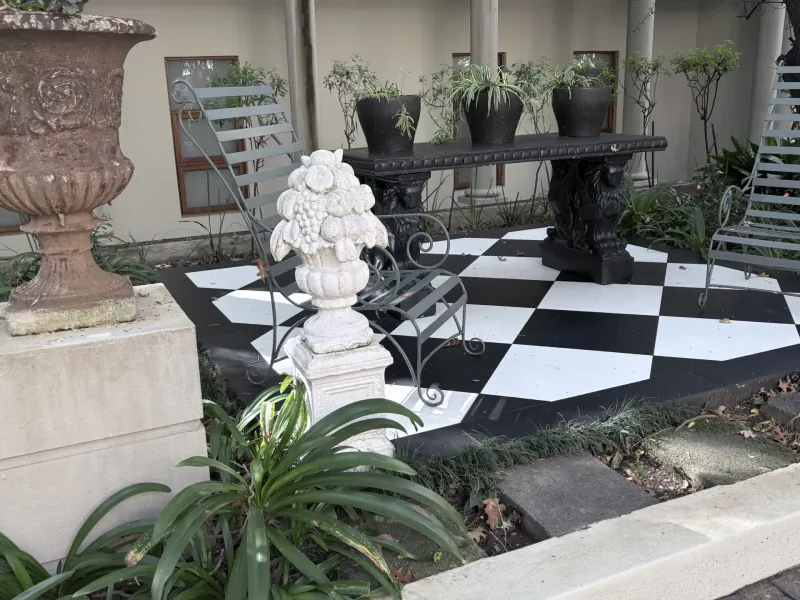 Outdoor patio with black and white checkered floor, ornate metal chairs, potted plants, and a white stone urn with grapes sculpture