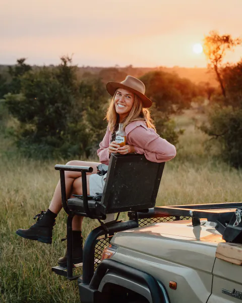 Woman in pink hoodie and hat smiling while holding a beer bottle on a safari vehicle at sunset