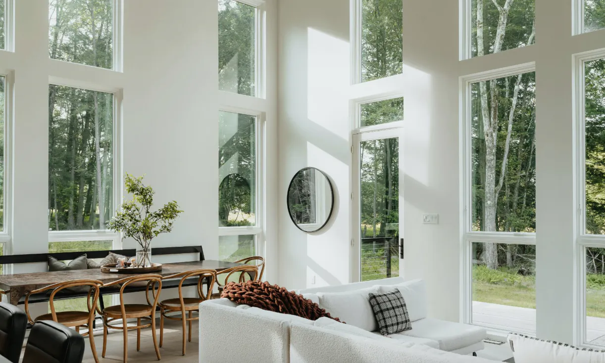 Bright modern living and dining room with white walls, large windows overlooking trees, white sofa, and wooden dining table