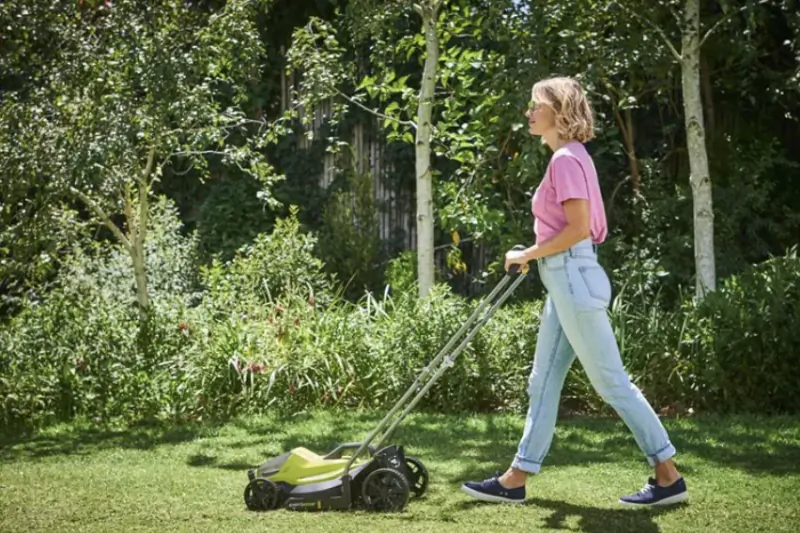 Woman in pink shirt pushing a green Ryobi cordless lawn mower across a sunny backyard lawn