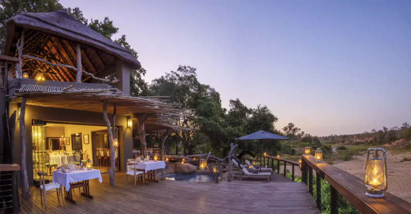 Twilight view of Savanna Stalkers' wooden lodge deck with dining tables, lounge chairs, pool, lanterns, and bushveld backdrop.