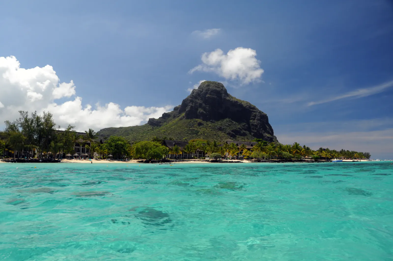 Turquoise ocean water with a tropical beach, palm trees, and a large rocky mountain under a blue sky with clouds