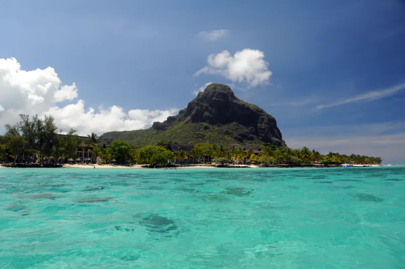 Turquoise ocean water with a tropical beach, palm trees, and a large rocky mountain under a blue sky with clouds