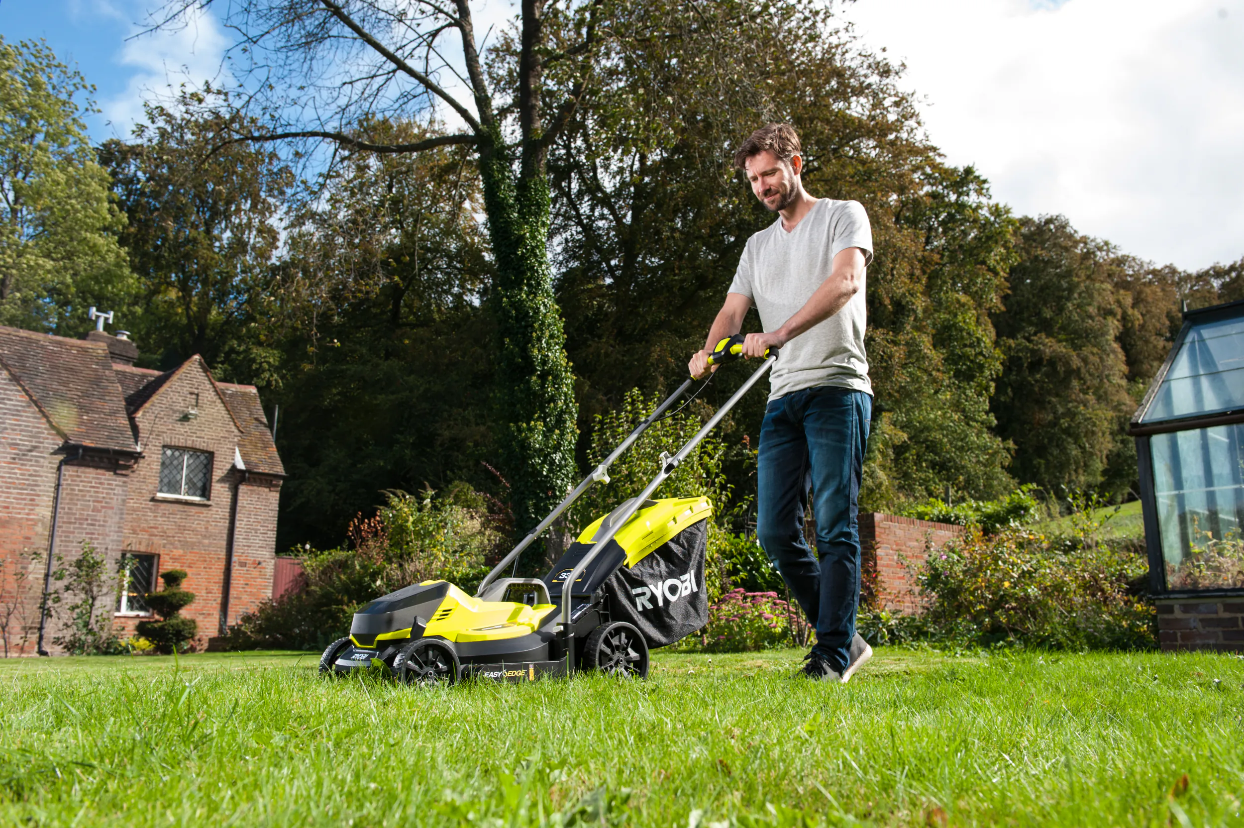 Man pushing a bright yellow and black Ryobi lawnmower across a green lawn in front of a brick house