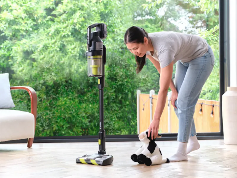 Woman in grey t-shirt and jeans bending to pick up a black-and-white stuffed cow beside a Ryobi cordless vacuum cleaner in a bright living room