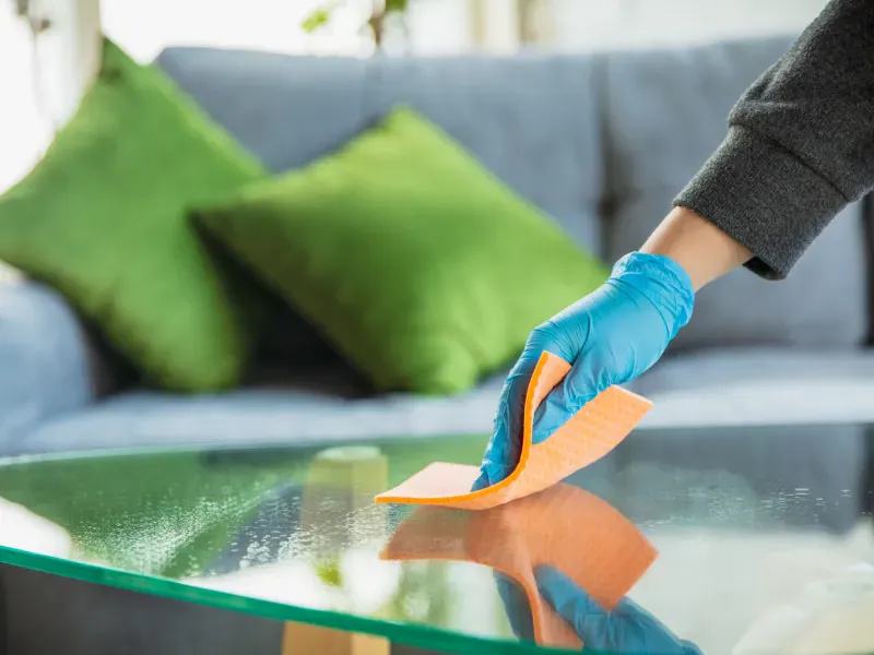 Gloved hand wiping a glass table with an orange cloth in a living room