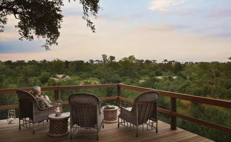 Woman relaxing in wicker chair on wooden deck overlooking African savanna bush at sunset with lanterns and trees.