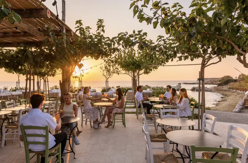 Groups of people dining at sunset on outdoor terrace at Cap St Georges Hotel & Resort, Akamas, with sea view and grapevine pergola.