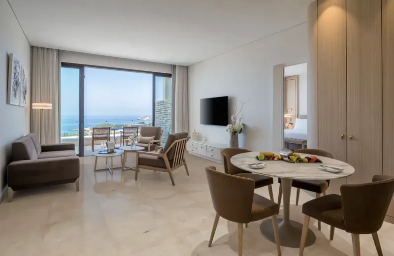 Luxurious hotel suite with brown leather sofas, round dining table with fruit, and panoramic sea view balcony at Cap St Georges Resort.