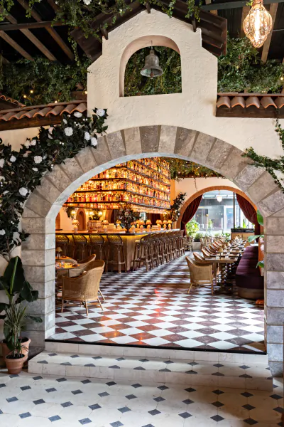 View through a stone archway into a vibrant restaurant with a checkered floor, bar, and hanging plants