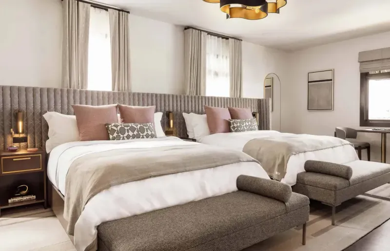 Modern hotel bedroom with two beds, beige and pink pillows, gray benches, and large windows with curtains