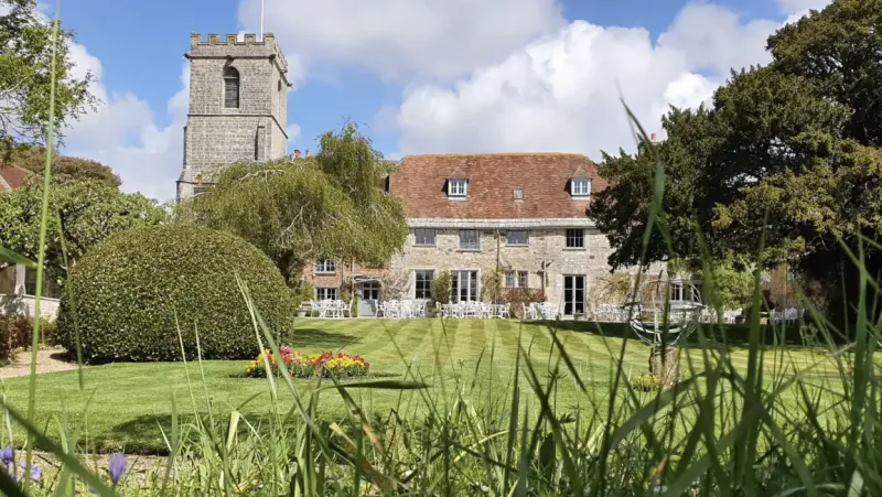 Stone manor house with red tiled roof and adjacent church tower on a sunny lawn
