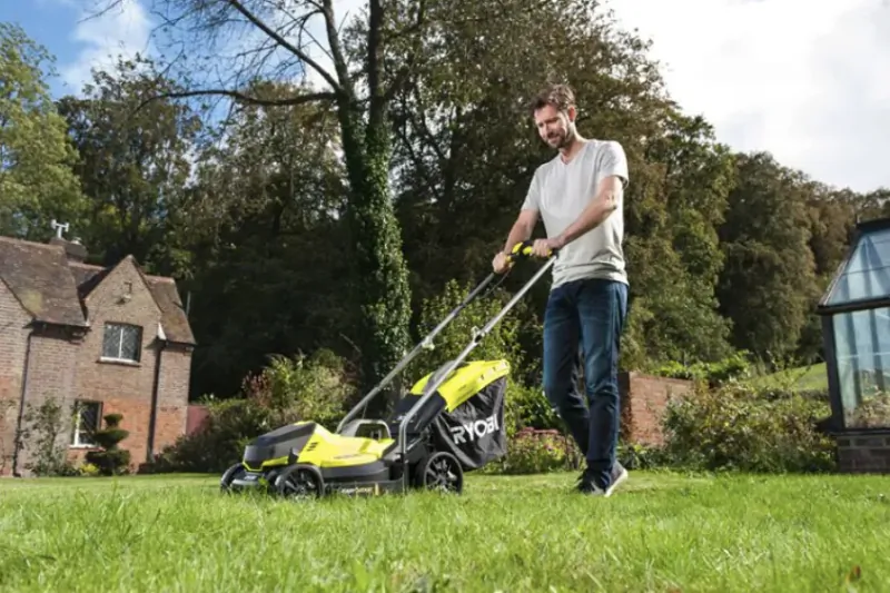 Man pushing a bright yellow and black Ryobi cordless lawn mower across a green lawn in front of a brick house