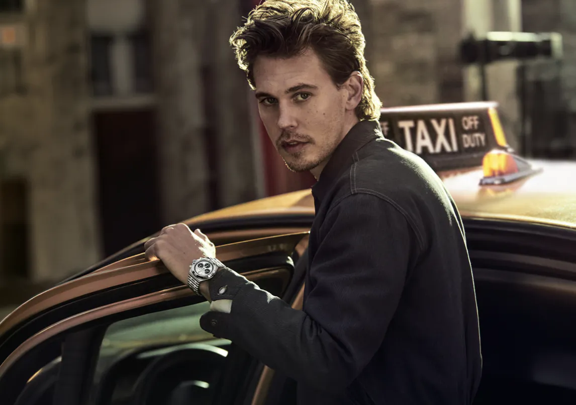 Man in dark jacket leaning on open door of yellow taxi, wearing silver watch, looking at camera