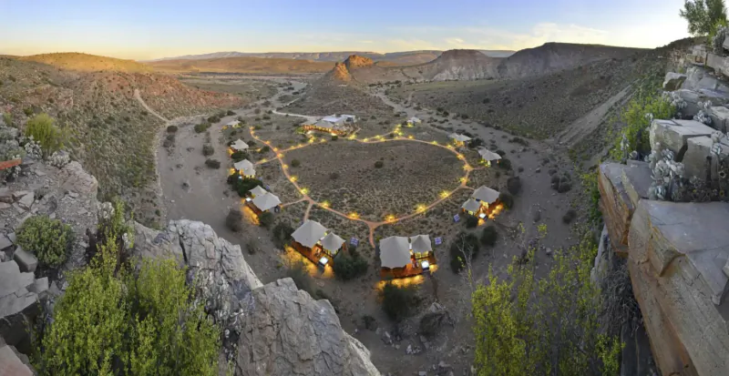 Aerial view of heart-shaped cluster of lit safari tents in African bush valley at sunset.