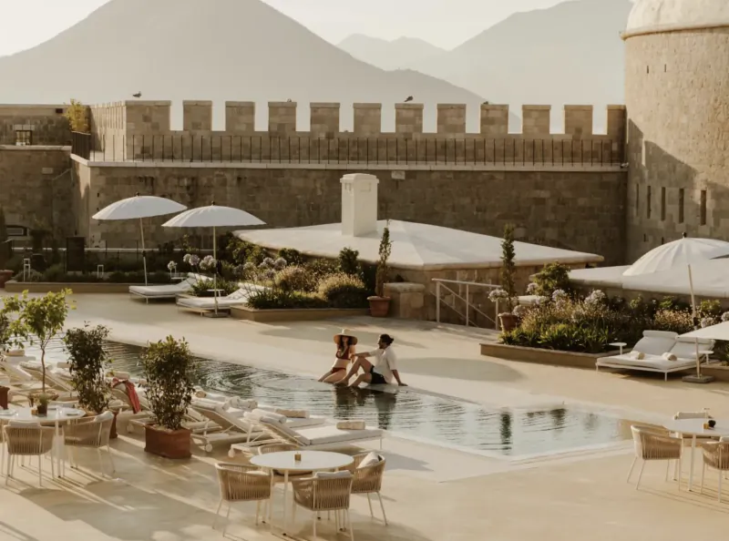 Two people sitting at the edge of a pool at a luxury resort with lounge chairs, umbrellas, and stone fortress walls with mountains in the background
