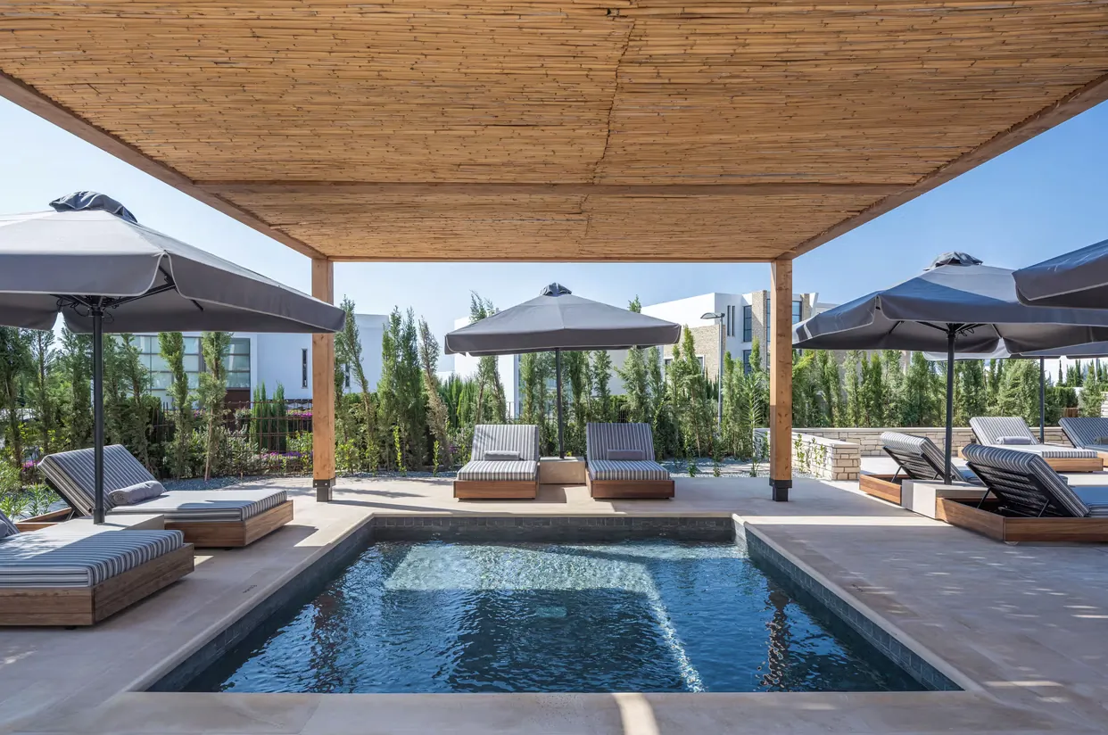 Luxury resort infinity pool with gray umbrellas, cushioned loungers, bamboo pergola, and Akamas sea view.