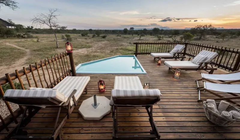 Infinity pool and white loungers on wooden deck overlooking African savanna at sunset with lanterns.