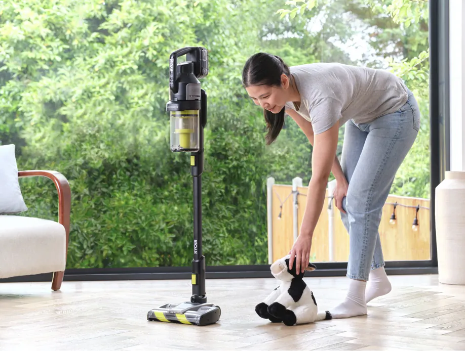 Woman bending to vacuum a stuffed cow toy with a Ryobi cordless stick vacuum in a bright living room
