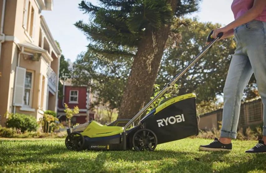 Person pushing a Ryobi cordless lawn mower across a green lawn in front of a house