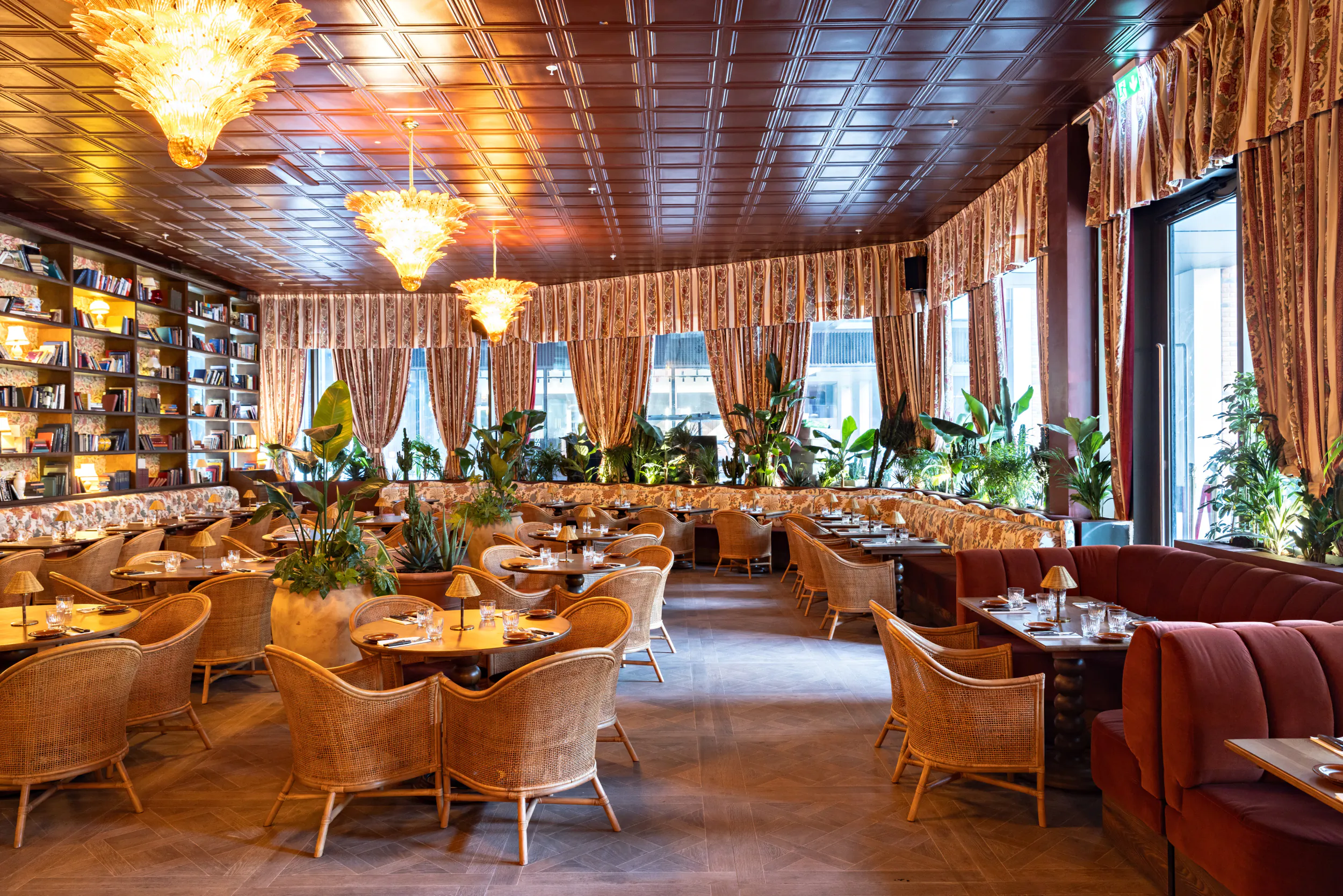 Elegant restaurant interior with wicker chairs, red velvet booths, bookshelves, and hanging chandeliers